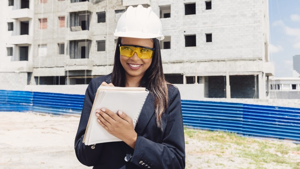 heureuse femme afro americaine dans un casque de securite ecrit dans un cahier pres du batiment en construction